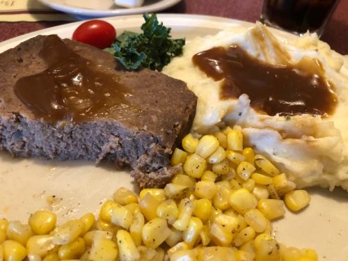 A plate with meatloaf topped with gravy, mashed potatoes, corn, and a cherry tomato on the side.
