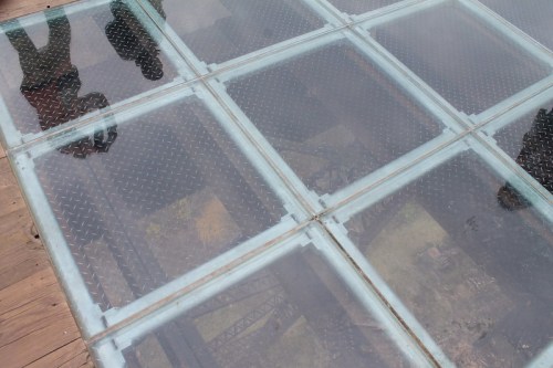 A glass floor panel showing reflections and a view of the ground below, with two people standing on it.