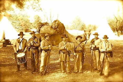 A group of soldiers in historical uniforms poses with rifles near a camel and tents in a sepia-toned setting.
