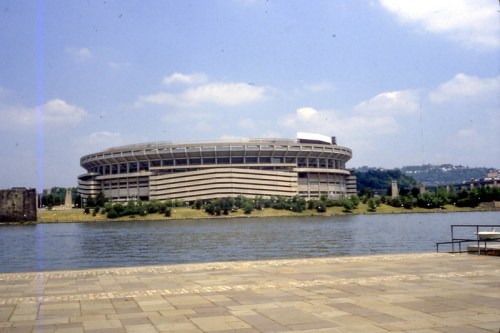 A large stadium by a river, surrounded by greenery and under a blue sky with scattered clouds.