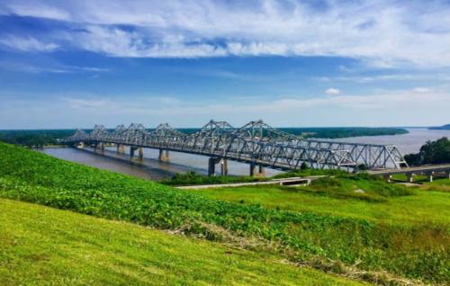 A large metal bridge spans a river, surrounded by green grass and trees under a blue sky with scattered clouds.