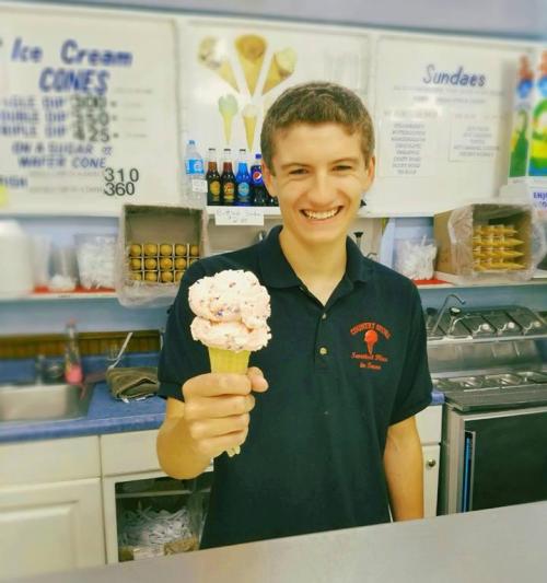 A smiling young man in a black shirt holds a cone of pink ice cream at an ice cream shop.