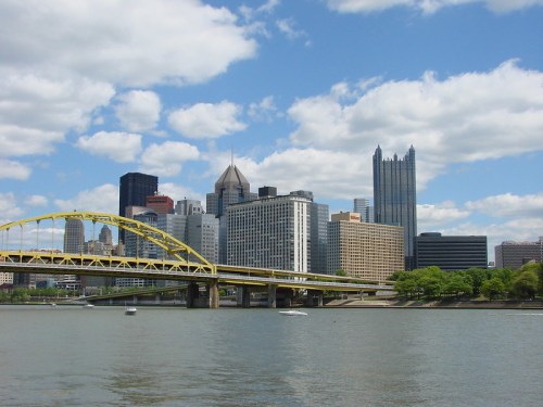 A view of Pittsburgh's skyline featuring a yellow bridge over a river, with clouds in a blue sky above.
