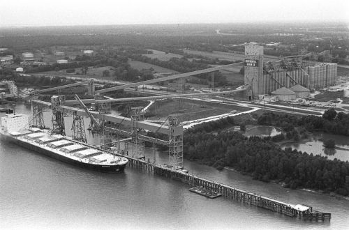 Aerial view of a grain elevator and loading dock with a cargo ship docked alongside, surrounded by fields and trees.