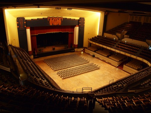 A spacious auditorium with empty rows of chairs, stage lights dimmed, and a vintage art deco design.
