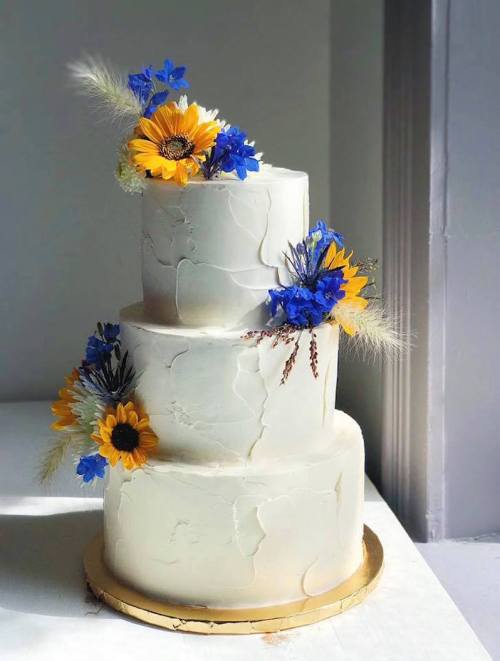 A three-tiered white cake adorned with sunflowers and blue flowers, featuring a textured finish and a gold base.