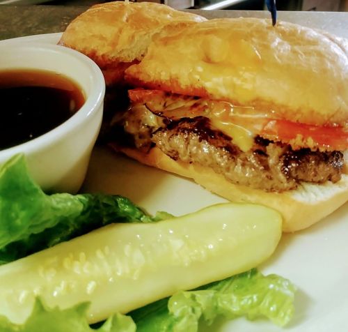 A close-up of a burger with cheese and tomato on a bun, served with a side of dipping sauce, lettuce, and a pickle.