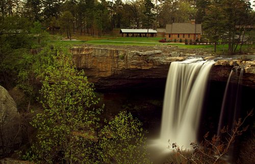 A serene waterfall cascades over a rocky ledge, surrounded by lush greenery and a rustic building in the background.