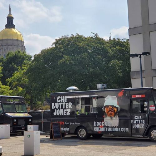 Food trucks parked in a city area, with a dome building in the background and trees providing shade.