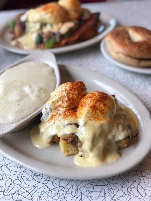A plate of eggs Benedict with creamy sauce, served over biscuits, alongside a dish of grits and a bagel in the background.