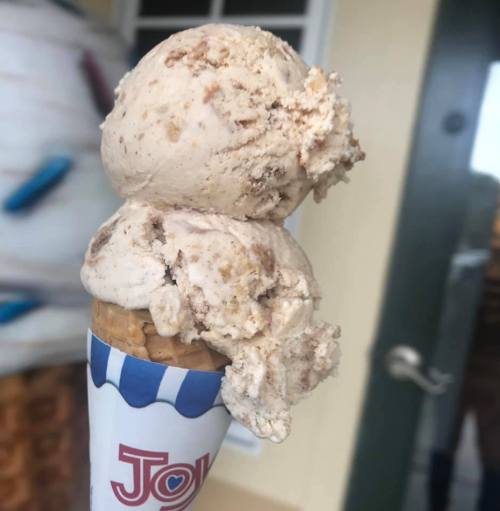 A close-up of a two-scoop ice cream cone with a creamy, light brown flavor, topped with a sprinkle of texture.