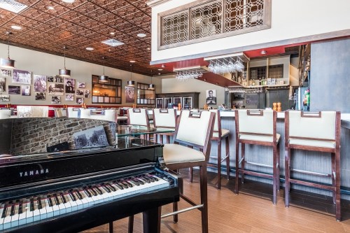 A stylish bar with a piano in the foreground, featuring wooden stools and framed photos on the walls.