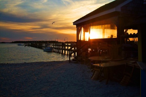 Sunset over a beachside restaurant with a pier, silhouetted tables, and a serene water view.