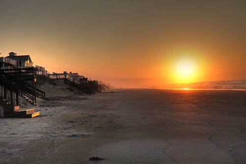 Sunset over a beach with wooden walkways and houses, casting a warm glow on the sand and ocean.