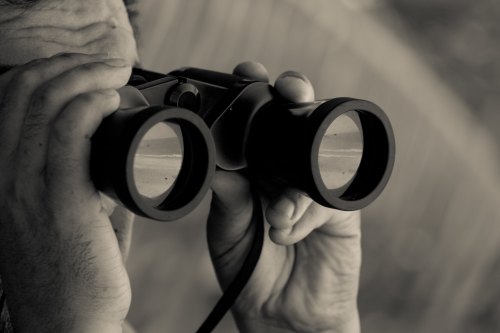 A person holding binoculars, looking through them with a blurred background. The image is in black and white.