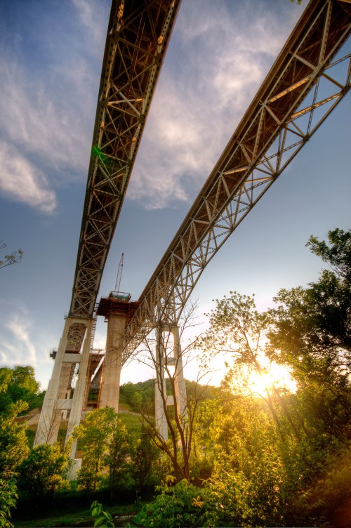 A large bridge under construction, with steel beams and supports, surrounded by trees and a sunset in the background.