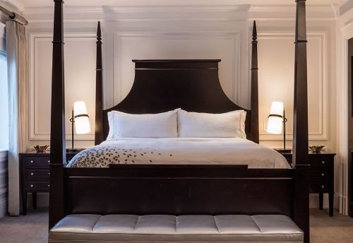 Elegant bedroom featuring a four-poster bed, white linens, and stylish bedside lamps against a neutral wall.
