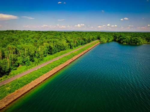 Aerial view of a serene lake bordered by lush green trees and a winding road along the shoreline under a blue sky.