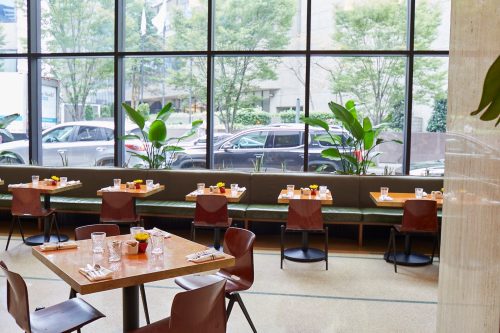 A modern restaurant interior with wooden tables, chairs, and large windows overlooking greenery and parked cars outside.