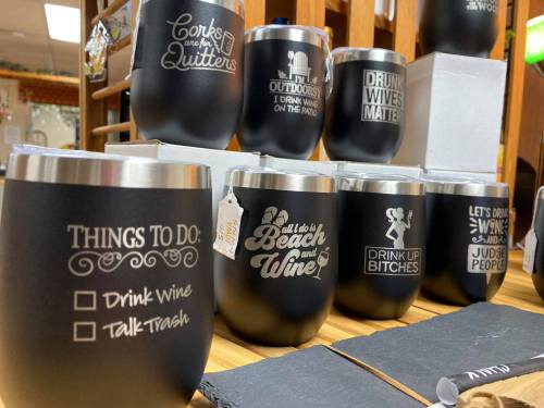 A display of black insulated wine tumblers with humorous phrases and to-do lists, arranged on a wooden shelf.
