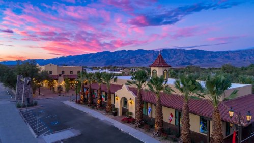 A scenic view of a building with palm trees at sunset, featuring colorful clouds and mountains in the background.