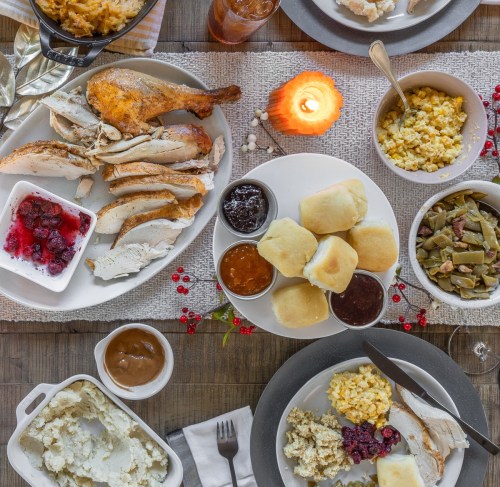 A festive table spread with roasted chicken, sides, rolls, and sauces, illuminated by a candle.