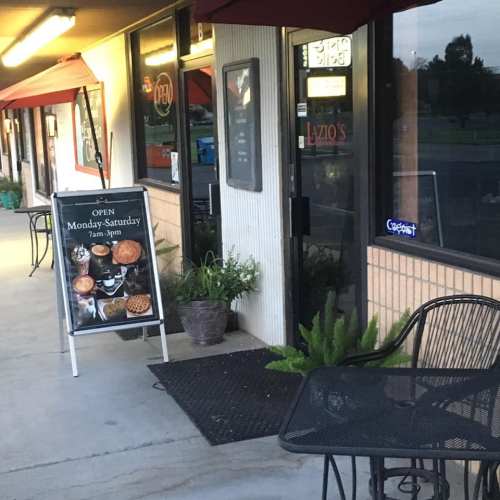 A storefront with a sign advertising hours and menu items, featuring outdoor seating and potted plants.