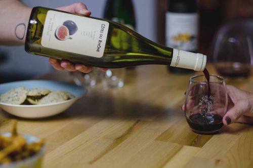 A hand pours red wine from a bottle into a glass, with snacks on a wooden table in the background.