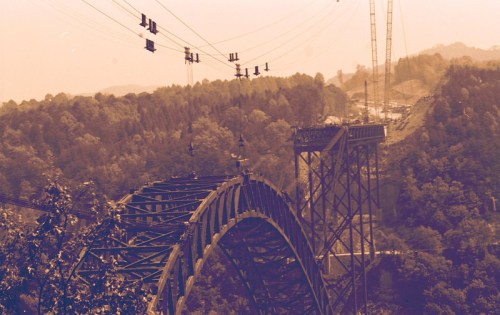 A vintage-style photo of a large metal bridge with cables, surrounded by trees and hills in the background.