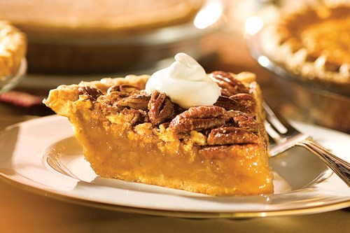 A slice of pecan pie topped with whipped cream on a white plate, with more pies in the background.