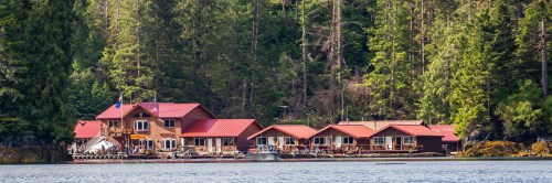 A scenic view of a wooden lodge with red roofs, surrounded by lush green trees and calm water.