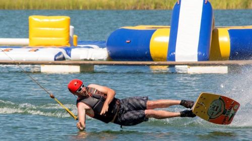 A person in a helmet and life vest is wakeboarding, leaning back as they glide over the water near inflatable structures.