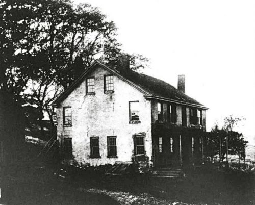 Black and white image of an old, two-story house surrounded by trees, showing signs of wear and age.