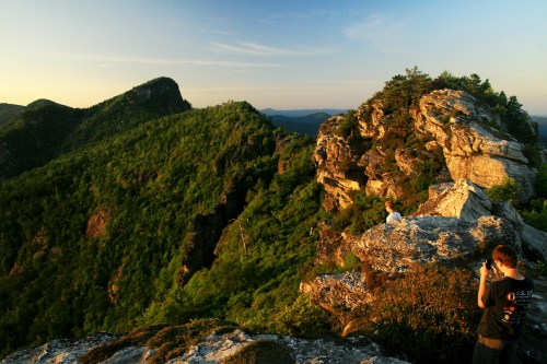Two hikers on rocky outcrops overlooking a lush green mountain landscape during sunset.