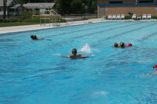 Children swimming in a large outdoor pool on a sunny day, with splashes and swim floats visible.