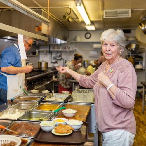 A smiling elderly woman gestures in a busy kitchen with various dishes and staff preparing food in the background.