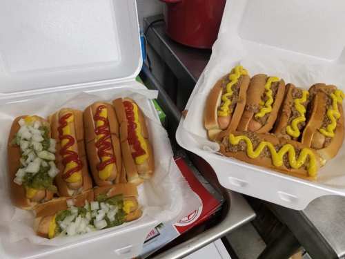 Two takeout containers filled with hot dogs, one topped with mustard, ketchup, onions, and pickles, the other with mustard and chili.