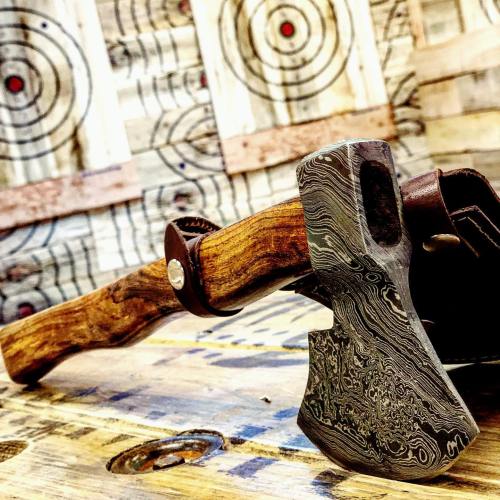 A close-up of a decorative axe with a patterned blade, resting on a wooden surface with target boards in the background.