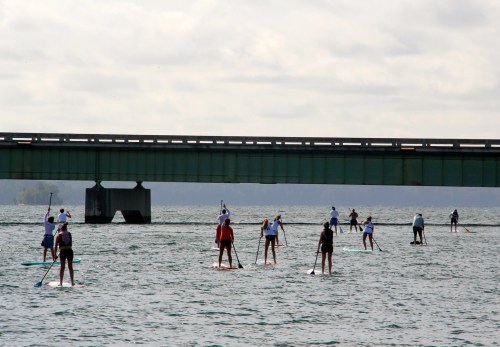 A group of people paddleboarding on a lake beneath a green bridge, with cloudy skies above.