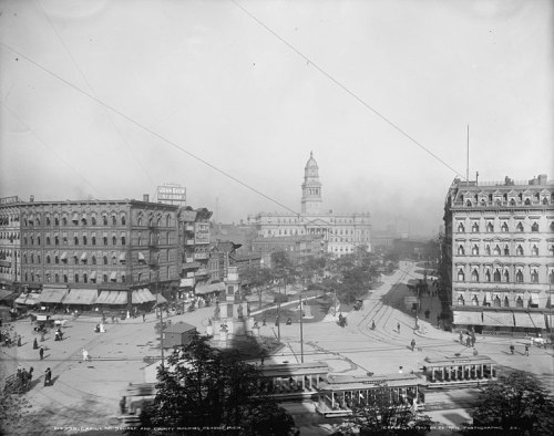 Historic view of a bustling city square with buildings, a monument, and people, captured in black and white.