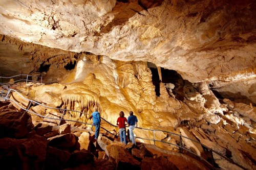 Three people explore a cave, admiring the rock formations and illuminated stalactites above.