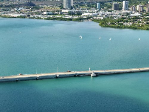 Aerial view of a bridge over turquoise water, with sailboats and a city skyline in the background.