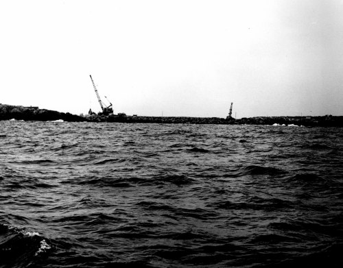 Black and white image of a body of water with cranes in the distance, set against a cloudy sky.
