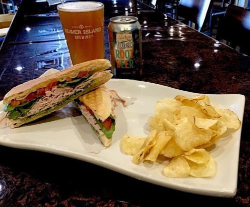 A sandwich with turkey, lettuce, and tomato, served with chips, alongside a beer and a can of diet root beer.