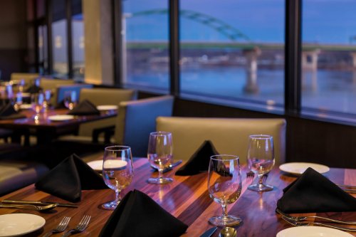 Elegant restaurant setting with tables set for dinner, featuring glassware and black napkins, overlooking a river view.
