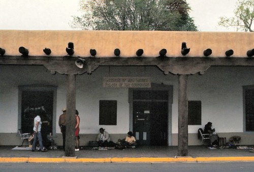A historic adobe building with wooden beams, featuring people sitting outside and a sign reading "Museum of New Mexico."