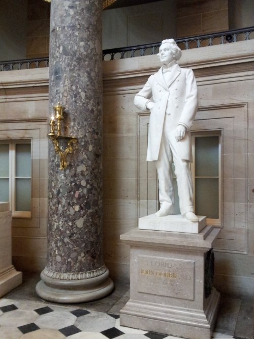 A white marble statue of a man in a coat stands beside a marble column in a grand interior space.