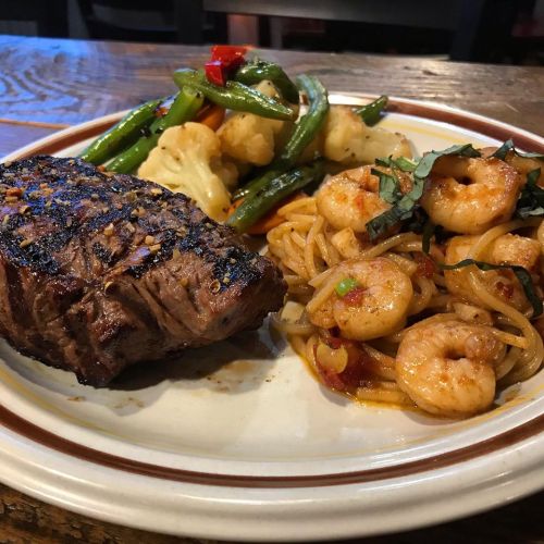 A plate featuring grilled steak, shrimp with noodles, and sautéed vegetables including cauliflower and bell peppers.