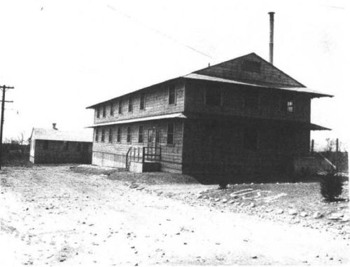 Black and white image of a two-story wooden building with a chimney, surrounded by dirt and gravel.