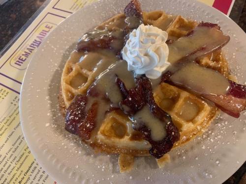 A waffle topped with crispy bacon, whipped cream, and drizzled with syrup, served on a white plate.